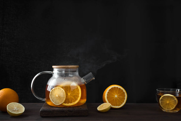 Citrus tea with  lemon, oranges  in transparent glass teapot on dark wooden table, copy space. Fruit tea. Seasonal winter autumn hot drink. - 写真・画像