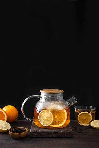 Citrusový čaj s citronem, pomeranče v moderní skleněné konvici na tmavém dřevěném stole, kopírovací prostor. Ovocný čaj. Sezónní zimní teplý nápoj. - Fotografie, Obrázek