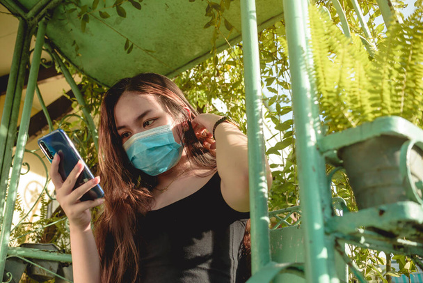 Una giovane donna asiatica che indossa una maschera per il viso controlla i suoi social media mentre si fissa i capelli. Si trova su un patio di metallo oscillare all'aperto. - Foto, immagini