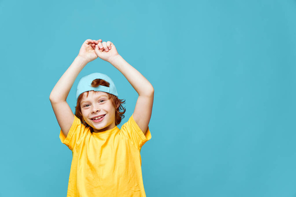 chico pelirrojo sosteniendo sus manos por encima de su cabeza azul gorra sonrisa amarillo camiseta  - Foto, Imagen
