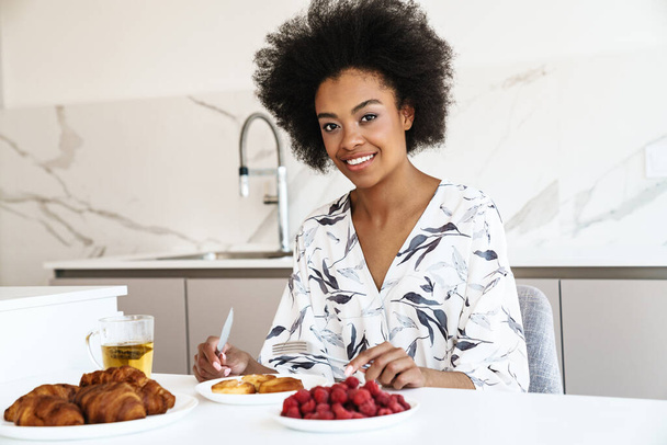 Cheerful beautiful woman having breakfast in kitchen, eating croissants and berries - Foto, imagen
