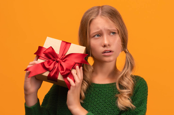 Image of charming blonde girl 12-14 years old in warm green sweater holding present box with red bow. Studio shot, yellow background, isolated. New Year Women's Day birthday holiday concept - Фото, изображение