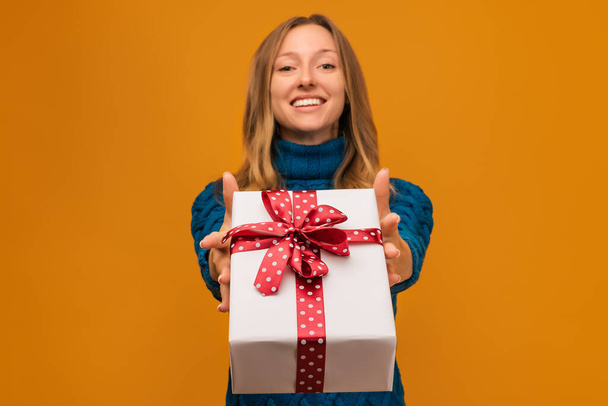 Cheerful young blonde woman in warm knitted sweater is showing present box to camera. Focus on gift box. Studio shot, yellow background. New Year Women's Day birthday holiday concept - Фото, изображение
