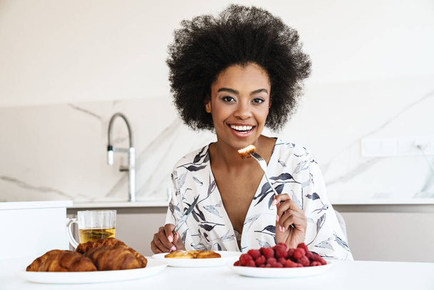 Cheerful beautiful woman having breakfast in kitchen, eating croissants and berries - Photo, image