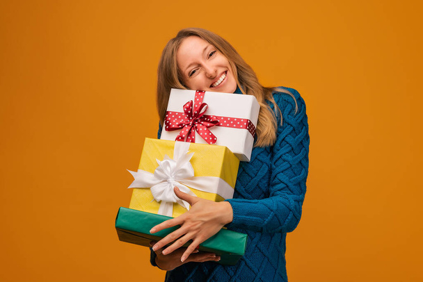 Charming young blonde woman in warm sweater holding a lot of gift boxes. Studio shot, yellow background. New Year, Women's Day, Birthday, Holiday concept - Photo, Image