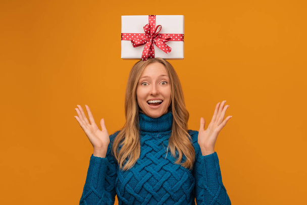 Charmante junge blonde Frau im Strickpullover hält Geschenkbox auf dem Kopf. Studioaufnahme, gelber Hintergrund. Neujahr, Frauentag, Geburtstag, Feiertagskonzept - Foto, Bild