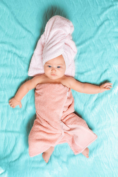 Ένα καυκάσιο μωρό τυλιγμένο σε μια ροζ πετσέτα ξαπλωμένο σε ένα μπλε σεντόνι με τα χέρια του απλωμένα σε διαφορετικές κατευθύνσεις. Θέα από ψηλά. Μωρό μετά το μπάνιο. - Φωτογραφία, εικόνα