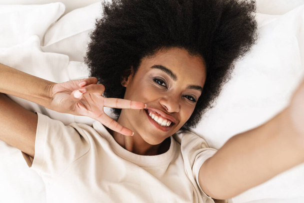 Beautiful african woman taking a selfie with smartphone while lying in bed isolated - Photo, image
