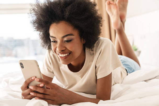 Young smiling african woman sending text messages, laying in bedroom - Photo, Image