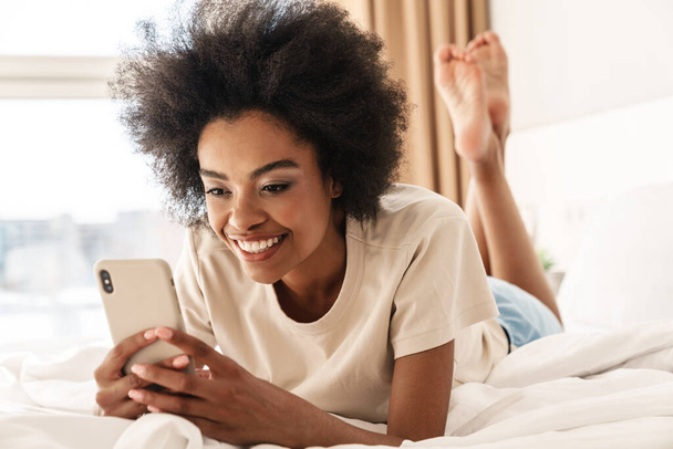 Young smiling african woman sending text messages, laying in bedroom - Foto, imagen