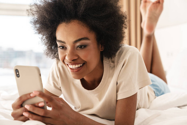 Image of beautiful cheerful african american girl smiling while using mobile phone in bad - Фото, зображення
