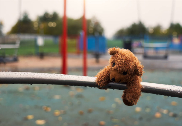 Χαμένη κούκλα αρκουδάκι ξαπλωμένη σε μέταλλο με σταγόνες βροχής στην παιδική χαρά σε ζοφερή μέρα, Μοναχική ή λυπημένη καφέ αρκούδα εγκαταλειφθεί ξαπλωμένη μόνη στο πάρκο, Μοναξιά έννοια, Διεθνής Ημέρα Αγνοουμένων Παιδιών - Φωτογραφία, εικόνα