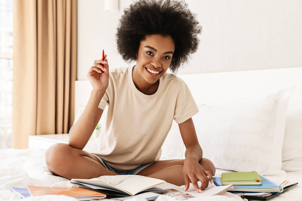 Smiling young african girl studying while sitting on bed with textbooks - Foto, Bild