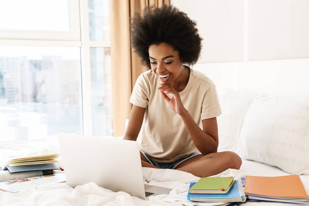 Smiling young african girl studying while sitting on bed with laptop computer - Foto, Bild