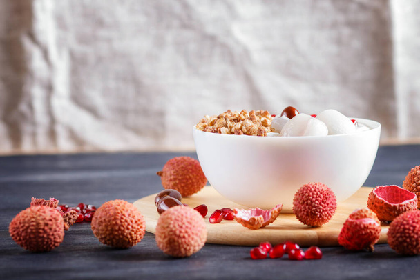 греческий йогурт с личи, гранатовые семена и мюсли в белой тарелке на черном деревянном фоне, закрыть. - Фото, изображение