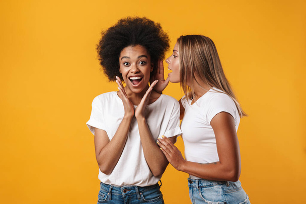 Photo of emotional happy young multiracial girls friends gossiping isolated over yellow wall background - Fotó, kép