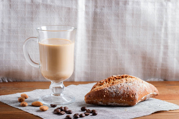 Glass cup of coffee with cream  and bun on a wooden  background and linen textile. close up, copy space. - Photo, Image