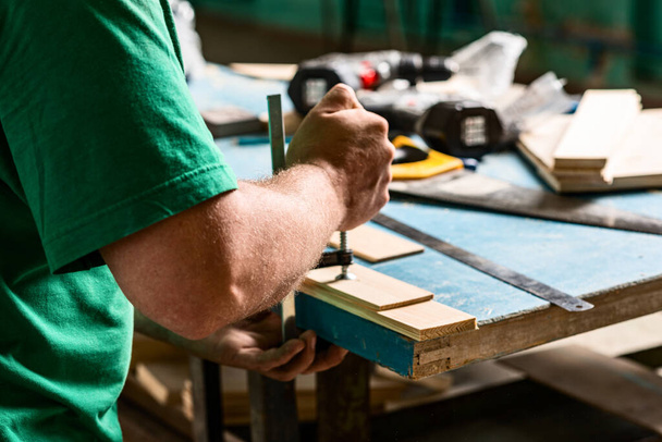 A man in a green t-shirt tightens the clamp on the table. Under the clamps are wood products. - Photo, Image