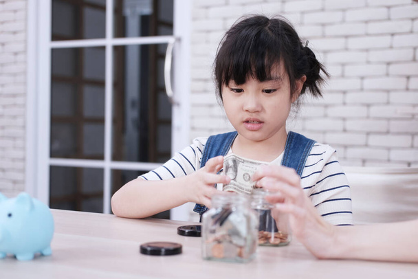 Uśmiechnięta azjatycka dziewczynka wkłada monety do skarbonki za oszczędność pieniędzy na drewnianym stole. Edukacja dla dzieci w ramach koncepcji wychowania domowego. - Zdjęcie, obraz