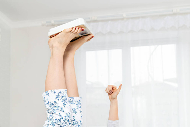 Eine Frau liegt mit erhobenen Beinen und auf ihnen liegenden elektronischen Waagen auf einem Bett. Hand mit erhobenen Daumen. Ansicht von unten. Das Konzept der Gewichtsabnahme und Gewichtszunahme. Füße und Hand aus nächster Nähe. - Foto, Bild