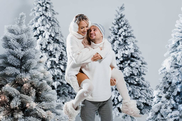 Пара поцелуев и объятий. Мужчина держит девушку возле рождественских деревьев в зимний день. Празднование Нового года. Люди в меховых наушниках, шляпах, белых свитерах. - Фото, изображение