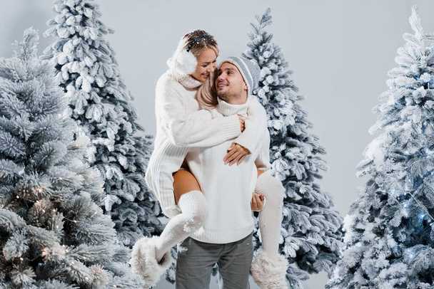 Couple kiss and hug. Man holds girl near christmas trees in winter day. New year celebration. People weared wearing fur headphones, hats, white sweaters. - Photo, image