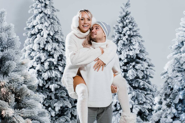 Couple kiss and hug. Man holds girl near christmas trees in winter day. New year celebration. People weared wearing fur headphones, hats, white sweaters. - Foto, Imagem