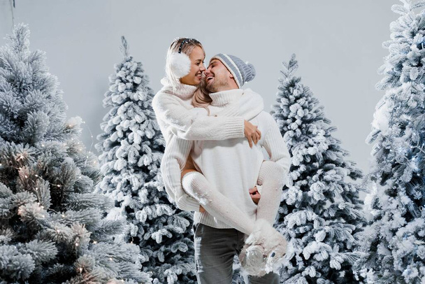 Couple kiss and hug. Man holds girl near christmas trees in winter day. New year celebration. People weared wearing fur headphones, hats, white sweaters. - Photo, Image