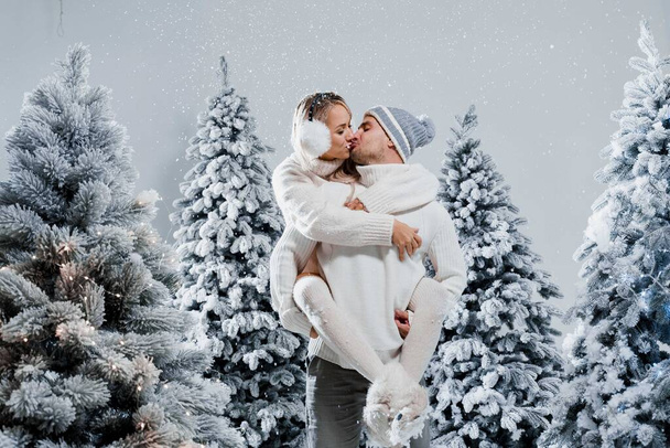 Ένα ζευγάρι φιλιά και αγκαλιές. Άντρας κρατά κορίτσι κοντά σε χριστουγεννιάτικα δέντρα την ημέρα του χειμώνα. Πρωτοχρονιά. Οι άνθρωποι φορούσαν γούνινα ακουστικά, καπέλα, λευκά πουλόβερ. - Φωτογραφία, εικόνα