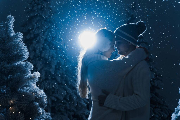 Падающий снег и поцелуи с падающим снегом и темно-синим фоном. Счастливая молодая пара носила белые пуловеры. обнимает и целует перед рождественскими деревьями в канун Нового года в зимний день - Фото, изображение