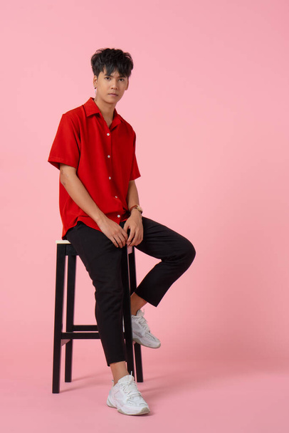 портрет молодого красивого хипстера азиатского мужчину в красной рубашке, сидящего на стуле, розовый фон - Фото, изображение