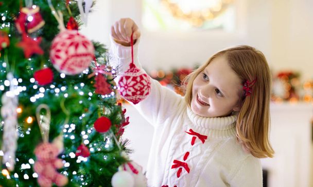 自宅でクリスマスツリーを飾る子供。Xmasの装飾が施されたニットセーターの少女。子供たちと家族は冬の休日を祝います。子供たちはクリスマスのためにリビングルームと暖炉を飾る. - 写真・画像