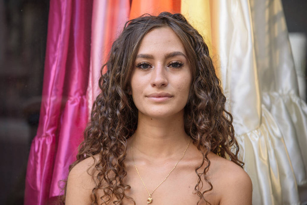 Close-up face and bare shoulders of beautiful young girl. Smiling woman with curly brown hair. Background with multicolored clothes. - Photo, Image