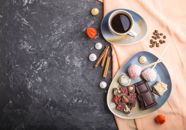 A pieces of homemade chocolate with coconut candies and a cup of coffee on a black concrete background and orange textile. top view, flat lay, copy space. - Fotoğraf, Görsel