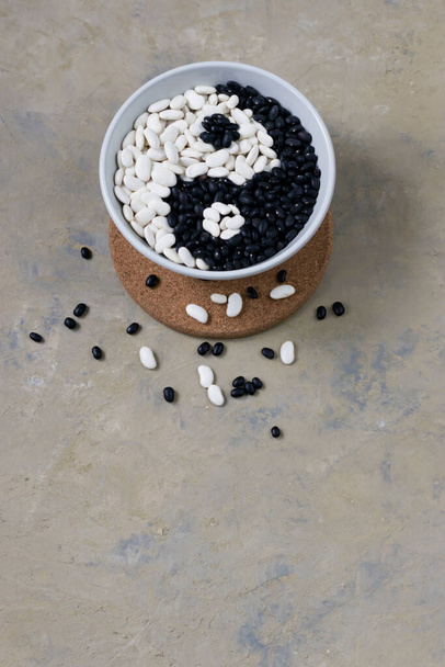 Legumes on a gray background. White bean and black bean in a light gray bowl on a wooden board. Sign Yin Yang. Vertical position. View up. Copy space - Fotoğraf, Görsel