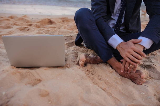 πλαϊνή άποψη ενός νεαρού άνδρα με κοστούμι και φορητό υπολογιστή που κάθεται στην παραλία και εργάζεται στην παραλία - Φωτογραφία, εικόνα