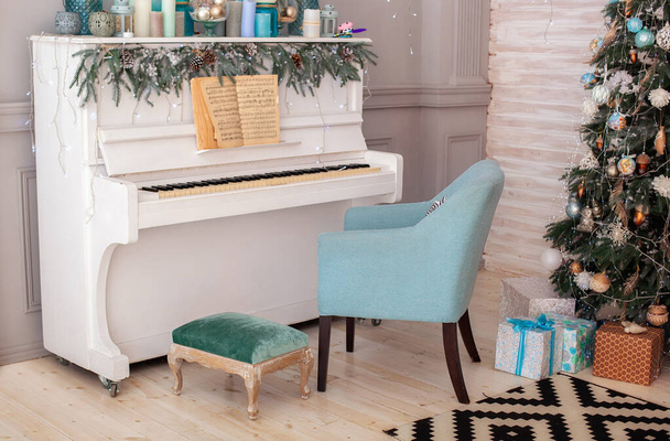 Sala de estar interior é decorada em estilo clássico com piano branco, árvore de Natal e presentes. Decoração de Ano Novo. Piano no quarto velas decoradas para o Natal. Piano com notas e poltrona azul - Foto, Imagem