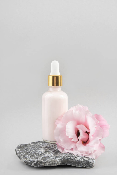 Anti-aging collagen, facial serum or moisturizing cream in glass bottle with pipette on stone and pink flower against grey background. Natural Organic Spa Cosmetic Beauty Concept Front view. - Fotografie, Obrázek
