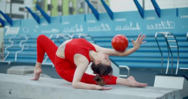 Жінка в червоному спортивному костюмі, що виконує телепередачі з гімнастичним м'ячем на ігровому майданчику на відкритому повітрі, спорт у міському середовищі, гімнастика в місті, 4k 120p Prores HQ - Кадри, відео