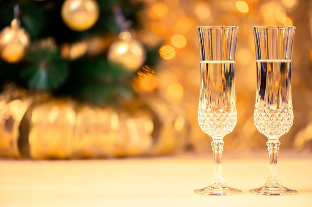Krásné sklenice na šampaňské na pozadí vánočního stromku a věnce v malé hloubce pole. Teplé osvětlení ve žlutých a zlatých tónech. - Fotografie, Obrázek