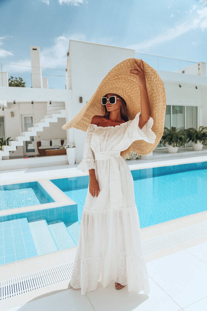 moda foto ao ar livre da bela mulher sexy com cabelo loiro em vestido branco luxuoso e grande chapéu de palha posando em villa branca perto da piscina - Foto, Imagem