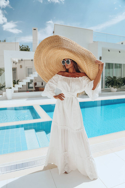 Havuza yakın beyaz bir villada poz veren güzel, sarışın, beyaz elbiseli ve büyük hasır şapkalı seksi bir kadının moda fotoğrafı. - Fotoğraf, Görsel