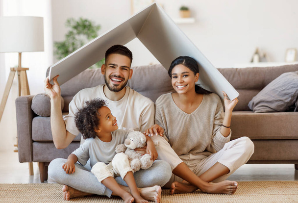 Χαρούμενοι γονείς με το παιδί χαμογελά και κρατώντας οροφή mockup πάνω από τα κεφάλια, ενώ κάθεται στο πάτωμα στο άνετο σαλόνι κατά τη διάρκεια relocatio - Φωτογραφία, εικόνα