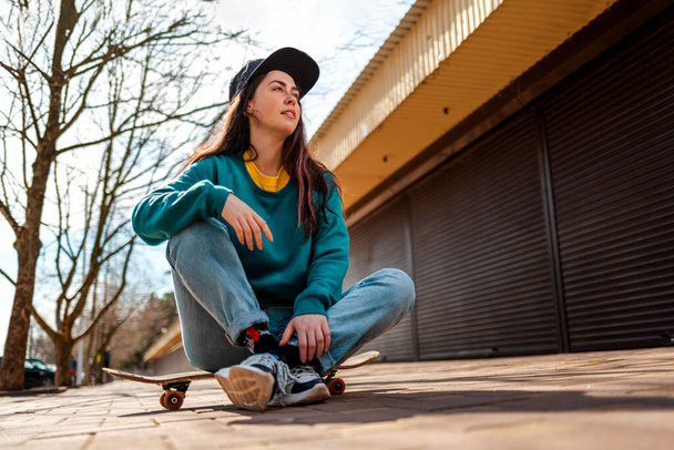Een jonge mooie blanke vrouw zit ontspannen op een skateboard en kijkt weg. Op de achtergrond, een straat met bomen. Onderaanzicht. Concept van sportieve levensstijl en straatcultuur. - Foto, afbeelding