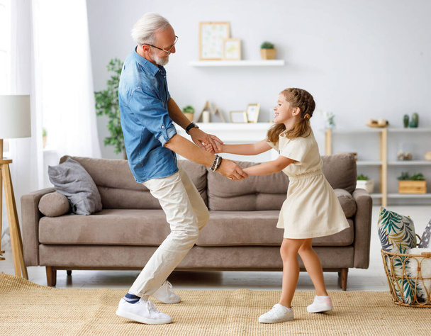 Happy family: girl smiling and dancing with aged grandfather while standing on sofa in cozy living room at hom - Foto, Imagen
