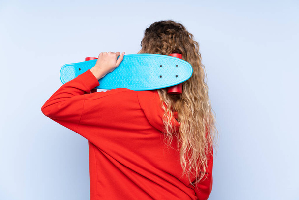 Young blonde woman with curly hair isolated on blue background with a skate in back position - Photo, Image