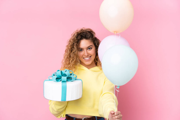 Young blonde woman with curly hair catching many balloons and holding a big cake isolated on pink background - 写真・画像