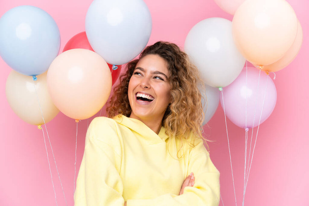 Young blonde woman with curly hair catching many balloons isolated on pink background - Фото, зображення