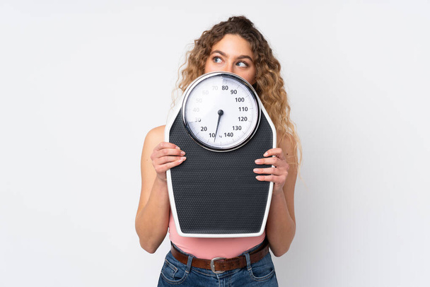 Young blonde woman with curly hair isolated on white background with weighing machine and hiding behind it - Foto, Imagen