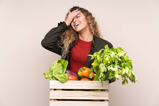 Фермер со свежесобранными овощами в коробке, изолированной на бежевом фоне смеясь - Фото, изображение
